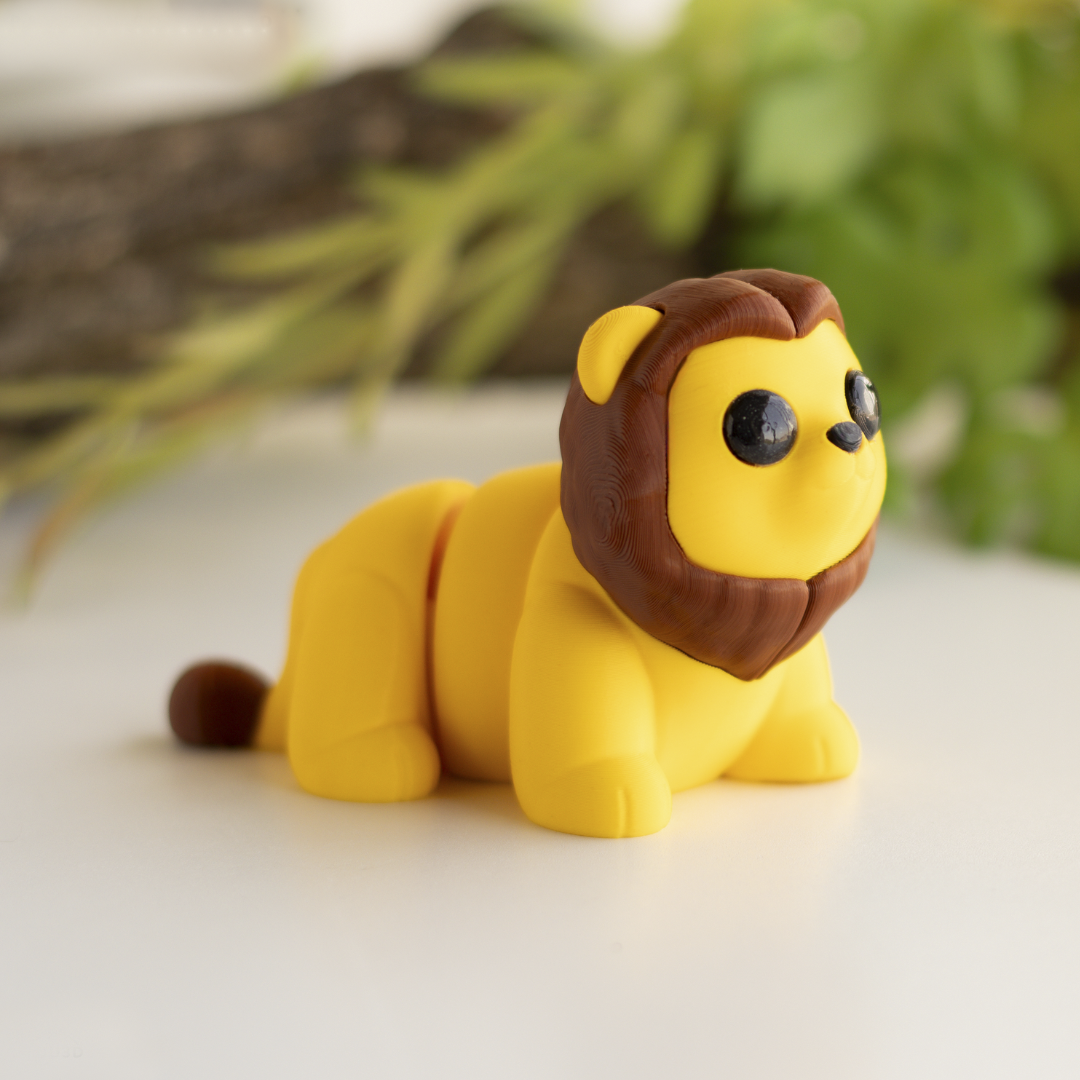A yellow toy lion with a brown mane and tail is sitting on a white surface, with green foliage in the background.