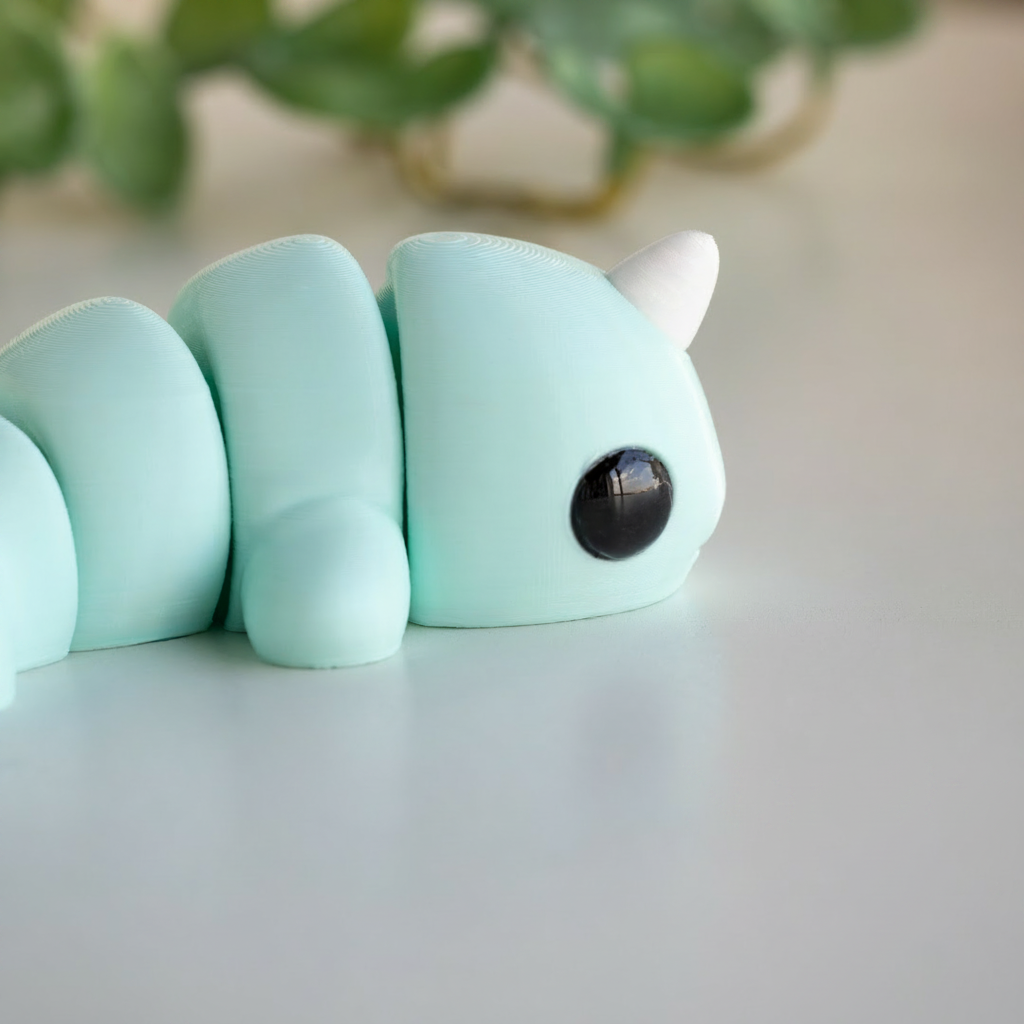 A light blue toy resembling a caterpillar or worm with a black eye and a white horn, sitting on a white surface.