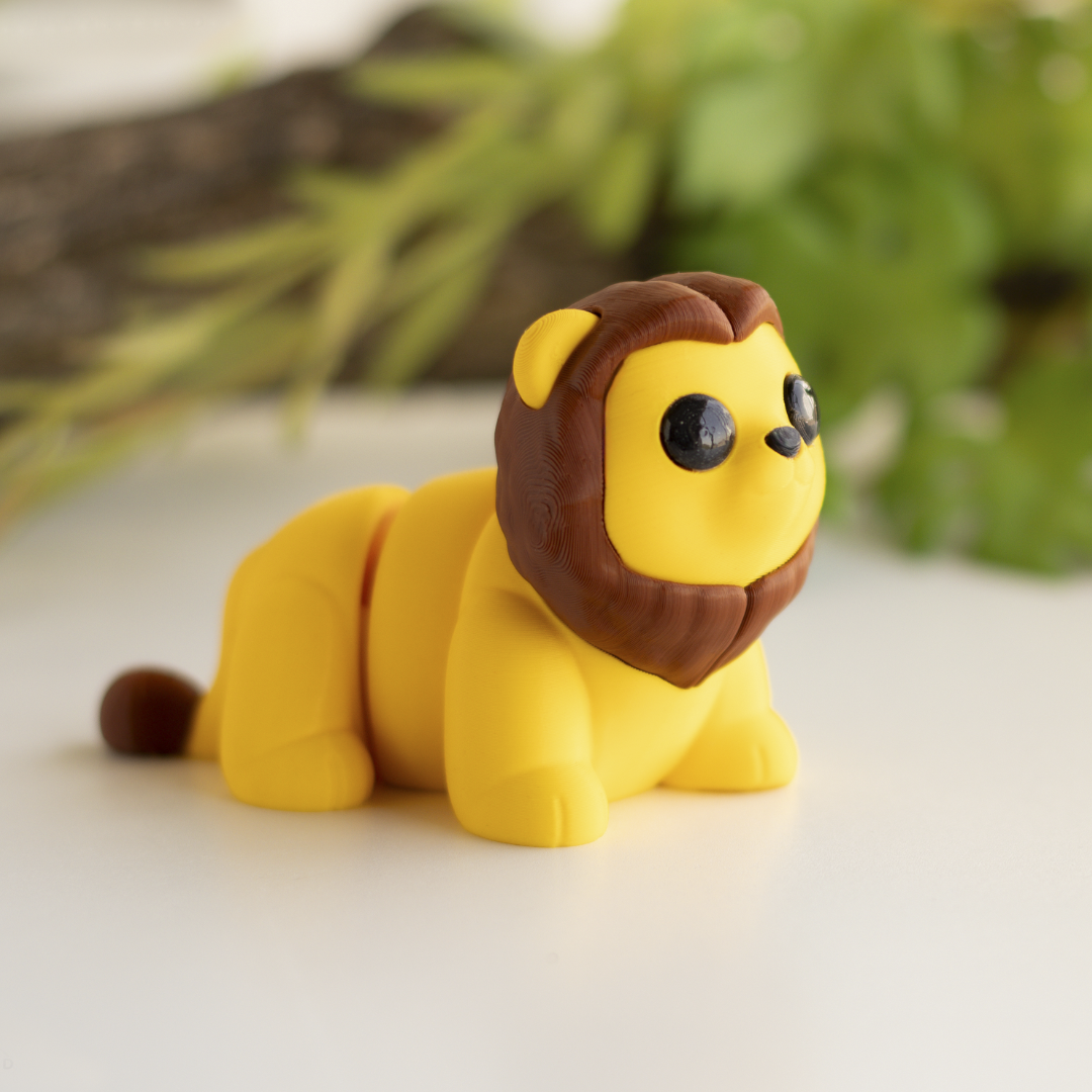 A yellow toy lion with a brown mane and tail is sitting on a white surface, with green foliage in the background.